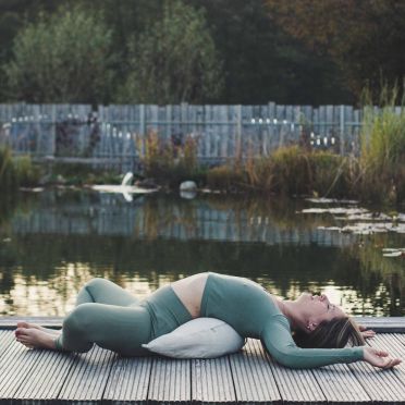 młoda kobieta w zielonym stroju do jogi leży na plecach na pomoście nad jeziorem , wsparta o jasną poduszkę do jogi i relaksacji w kształcie księżyca