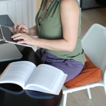 Kobieta przy biurku siedzi prosto na welurowej, pomarańczowej poduszce klinowej, obok leży książka i kubek herbaty.