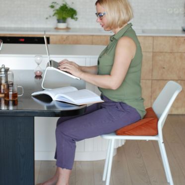 Kobieta pracuje na laptopie, siedząc na welurowej, pomarańczowej poduszce klinowej na białym krześle.