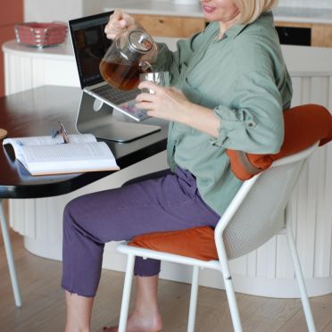 Kobieta siedzi przy stole na welurowej, pomarańczowej poduszce klinowej z wałkiem pod plecami, nalewając herbatę do szklanki.