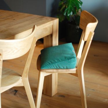 Zielona welurowa poduszka klinowa na drewnianym krześle przy stole, drewniana podłoga i roślina w tle.