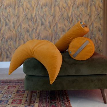 Poduszka w kształcie księżyca w jesiennym żółtym kolorze obok musztardowego bolstera do jogi, ułożone na zielonym fotelu, na tle wzorzystej tapety i orientalnego dywanu.