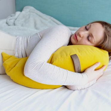 Młoda kobieta ubrana na biało w długich blond włosach leży na białej pościeli w pozycji na brzuchu, obejmując pod głową poduszkę medytacyjną w kształcie księżyca, w kolorze żółtym. w tle jasno niebieska ściana.Widok z boku.