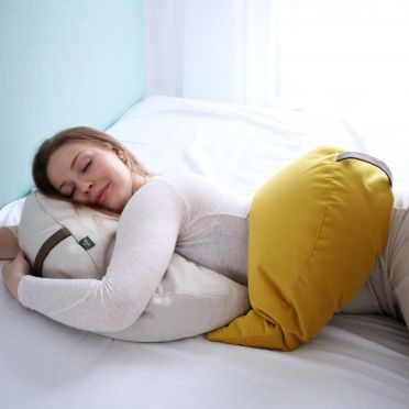 Młoda kobieta ubrana na biało w długich blond włosach leży na białej pościeli w pozycji na brzuchu, obejmując pod głową jasną poduszkę medytacyjną w kształcie księżyca, na biodrze na położoną taką samą poduszkę w kolorze żółtym. w tle jasno niebieska ściana.Widok z boku.