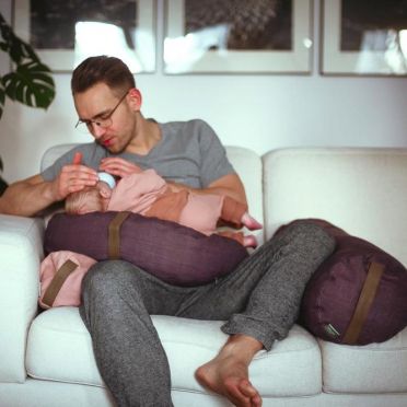 Mężyczna siedzący na kanapie karmi niemowlę z buteli. Niemowle leży na poduszce w kształcie księżyca na kolanach mężczyzny. noga mężczyzny wsparta jest o wałek bolster do jogi.