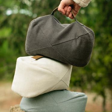 Trzy poduszki do medytacji w odcieniach grafitowym, beżowym i błękitnym ułożone w stos, na pniu drzewa; górna podnoszona ręką, w tle rozmyta zieleń lasu.