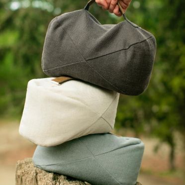 Trzy poduszki do medytacji w odcieniach grafitowym, beżowym i błękitnym ułożone w stos, na pniu drzewa; górna podnoszona ręką, w tle rozmyta zieleń lasu.