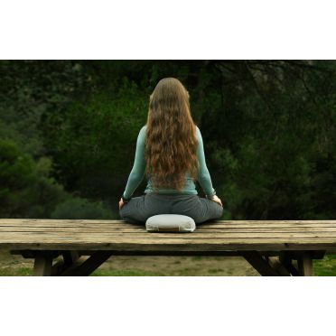 Kobieta z długimi, falowanymi włosami medytuje na szarej poduszce na drewnianym stole w otoczeniu zielonych drzew.