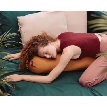 Kobieta w bordowym stroju leży w pozycji embrionalnej na macie do jogi, obejmując gryczany wałek do jogi z weluru w kolorze miodowo-musztardowym; w tle rośliny doniczkowe i przeszklenie z widokiem na patio.