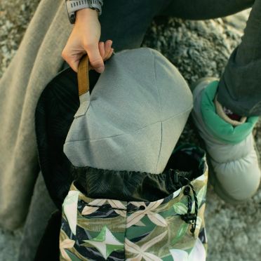 Zbliżenie na osobę wyciągającą jasnoszarą poduszkę do medytacji z kolorowego plecaka, w tle kamienne podłoże i zimowy strój sugerujący aktywność na świeżym powietrzu.