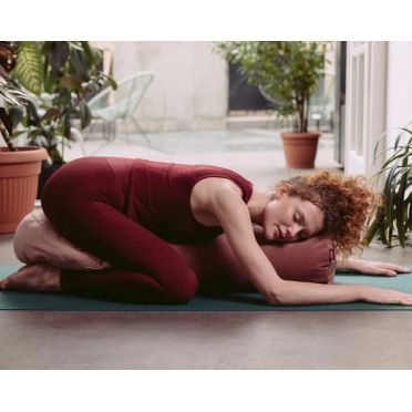 Kobieta w bordowym stroju leży w pozycji embrionalnej na macie do jogi, obejmując winno-różowy wałek WellB z wypełnieniem z łusek gryki; w tle rośliny doniczkowe i przeszklenie z widokiem na patio.
