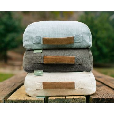 Trzy prostokątne poduszki do medytacji w kolorze błękitnym, grafitowym i beżowym, ułożone jedna na drugiej na drewnianej ławce w parku.