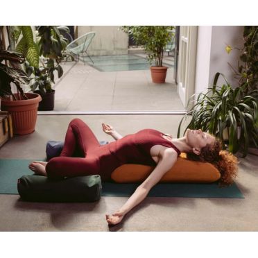 Kobieta w bordowym stroju leży w pozie relaksacyjnej na dużym wałku WellB w kolorze musztardowym, pod nogami ma dwa małe wałki gryczane.