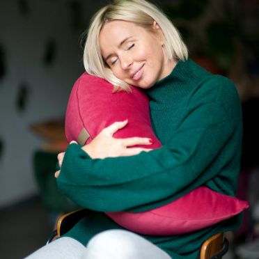 kobieta w blond włosach i zielonym swetrze przytula dużą poduszkę do medytacji i relaksacji w kolorze malinowego różu