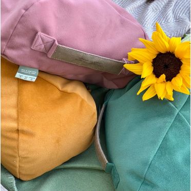 Trzy poduszki do medytacji w odcieniach różu, musztardowym i zielonym leżą na kocu; obok nich jasny słonecznik, w tle widoczny sweter i pled.