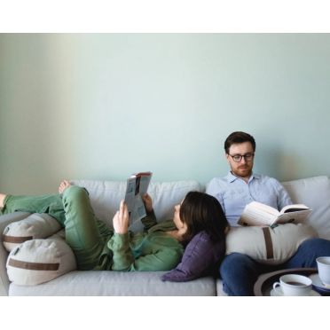 Para relaksuje się na jasnej sofie z książkami, używając piaskowych małych wałków gryczanych WellB jako podpórki pod nogi, głowę i na kolanach.