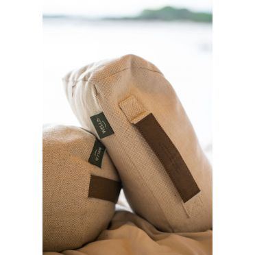 Zbliżenie na dwie beżowe poduszki do medytacji z zielonymi metkami WELLB i skórzanym uchwytem, ułożone na jasnym materiale, w tle rozmyty widok na wodę i drzewa.