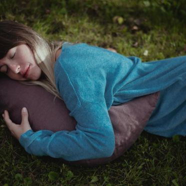 Kobieta w niebieskim swetrze leży na zielonej trawie, przytulając fioletowo-brązową poduszkę w kształcie półksiężyca, wyraźnie odprężona.