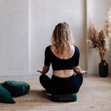 Kobieta w czarnym stroju medytuje w pozycji siedzącej na zielonej poduszce na plecionym dywanie; w tle jasna ściana z niszą i zielony wazon z suchymi trawami.