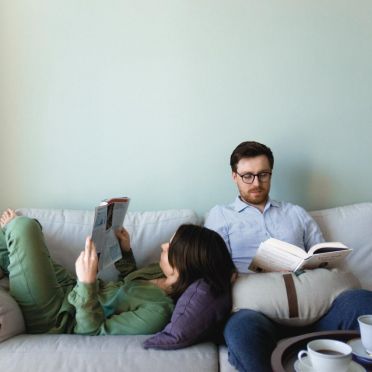 Kobieta i mężczyzna  odpoczywają na białej kanapie, mężczyzna siedzi i ma na kolanach  jasną poduszkę do relaksacji w kształcie księżyca. Kobieta leży na plecach, głowę ma wspartą o taką samą poduszkę w kolorze ciemnoszarym.