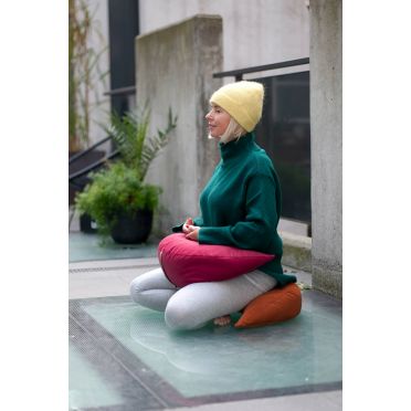 kobieta siedząca w pozycji medytacyjnej na poduszce do medytacji i relaksacji w kształcie księżyca w kolorze miedzianym, kobieta trzyma na podołku poduszkę w kształcie księżyca w kolorze malinowego różu.