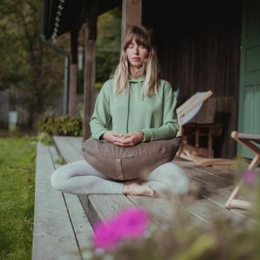Kobieta w zielonej bluzie medytuje na tarasie, siedząc na jasno-brązowej poduszce półksiężycowej WellB, w otoczeniu natury.