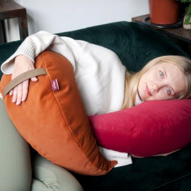 młoda kobieta leży na boki na ciemnozielonej kanapie, pod głową ma poduszkę medytacyjną w kształcie księżyca w kolorze malinowym, ciemnoróżowym, na biodrze na poduszkę w kolorze ruym, ciemnopomarańczowym.