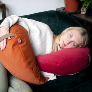 młoda kobieta leży na boki na ciemnozielonej kanapie, pod głową ma poduszkę medytacyjną w kształcie księżyca w kolorze malinowym, ciemnoróżowym, na biodrze na poduszkę w kolorze rudym, ciemnopomarańczowym.