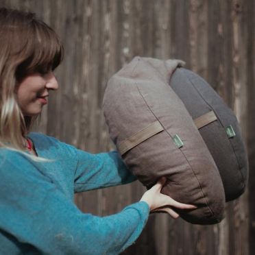 młoda kobieta w niebieskim swetrze trzyma w rękach dwie poduszki do medytacji i relaksacji w kształcie księżyca, w kolorach brązowym i grafitowym