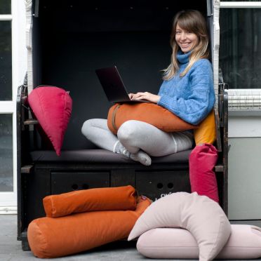 młoda dziewczyna siedzi na fotelu otoczona wieloma kolorowymi poduszkami medytacyjnymi z weluru, w różnych kształtach. Na kolanach trzyma laptop wsparty o poduszkę w kształcie księżyca w kolorze rudym, ciemnopomarańczowym. Pogodny nastrój zdjęcia.